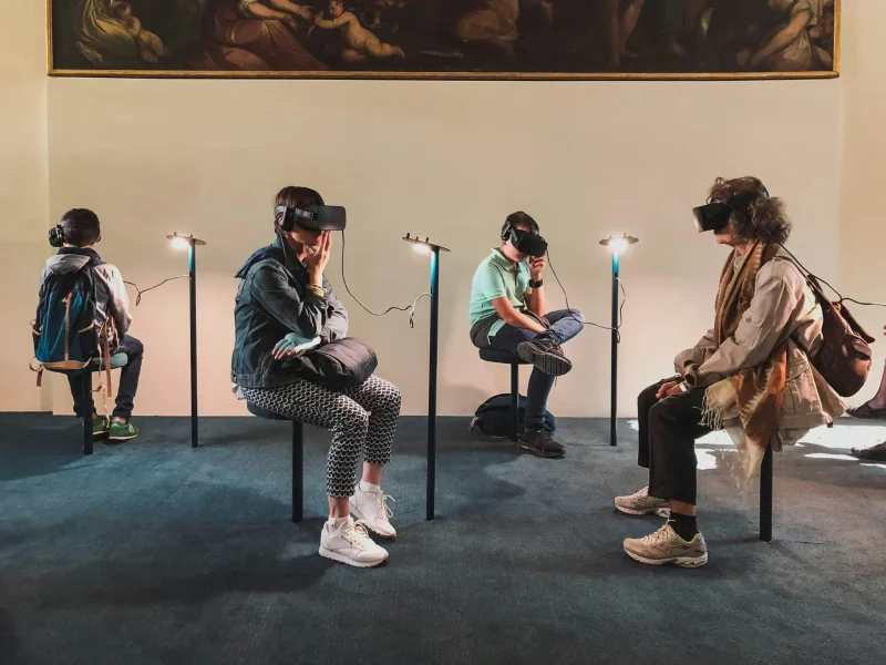Pengalaman VR dalam Pengaturan Galeri Empat orang yang memakai headset VR duduk di kursi, melihat sekeliling ruangan dengan lukisan di dinding.