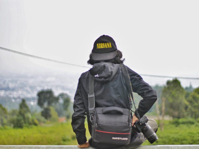 Seorang fotografer berdiri di tepi bukit dengan latar belakang pemandangan hijau dan kota yang kabur.