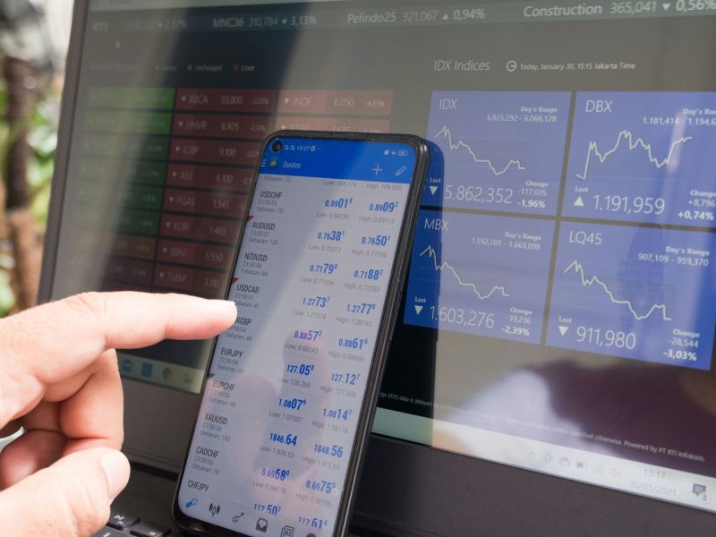 Data Keuangan pada Ponsel Pintar dan Laptop Gambar close-up tangan yang memegang ponsel pintar yang menampilkan data keuangan, dengan laptop di latar belakang yang menampilkan informasi serupa. Ponsel pintar menampilkan nilai tukar mata uang dan laptop menampilkan data pasar saham. Gambar tersebut menggambarkan hubungan antara kedua platform.