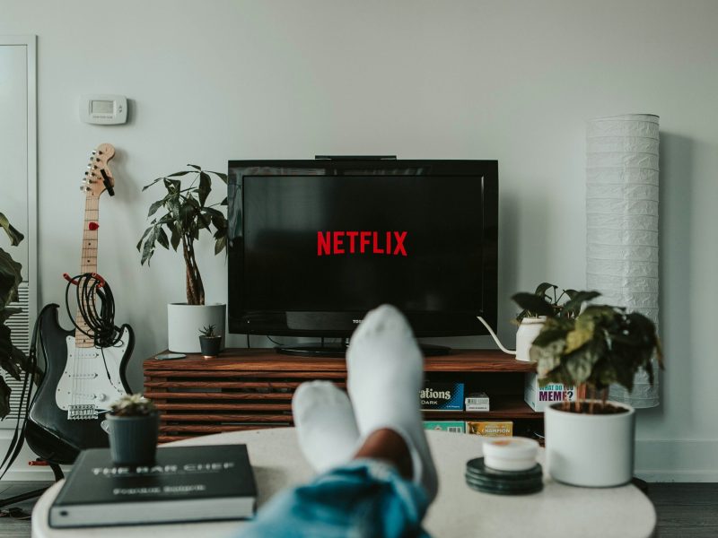 Seseorang bersantai di sofa dengan menonton Netflix, dikelilingi oleh tanaman dan alat musik.