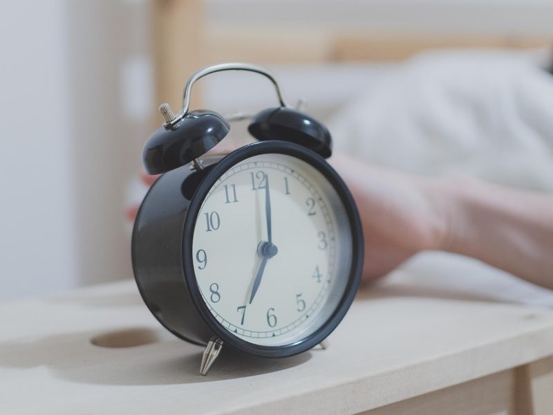 Jam alarm hitam klasik di atas meja kayu, dengan tangan seseorang di latar belakang.