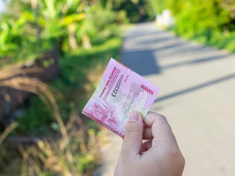 Uang Kertas Rupiah Tangan memegang uang kertas 100.000 Rupiah di depan latar belakang kabur berupa dedaunan hijau dan jalan beraspal.