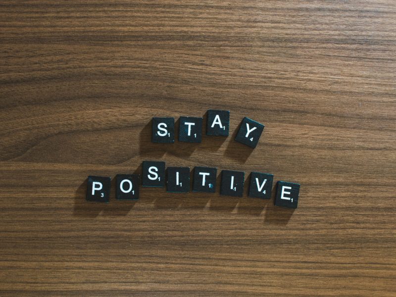 Pesan "Stay Positive" yang disusun dari huruf blok hitam di atas meja kayu.