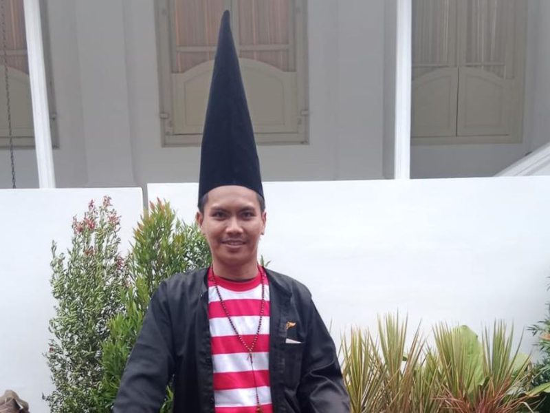 Pria mengenakan baju striped merah putih dan topi tinggi berdiri di depan taman.