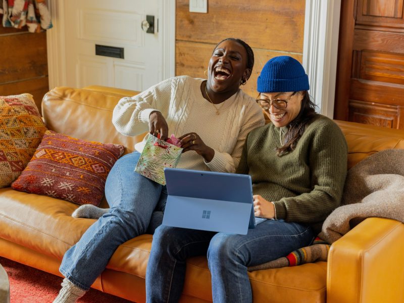 Dua Teman Tersenyum Dua orang sahabat, satu berkulit hitam dan satu Asia, duduk di sofa bersama sambil tertawa. Perempuan berkulit hitam itu memegang tas hadiah dan perempuan Asia itu sedang melihat laptop di pangkuannya. Mereka berdua mengenakan pakaian kasual dan sofa ditutupi selimut cokelat.