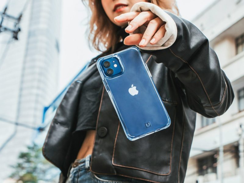 Wanita muda berpose dengan Iphone di tangannya sambil mengenakan jaket kulit hitam, kemeja putih, dan celana jeans biru. Latar belakang gambar menampilkan bangunan-bangunan di kota.