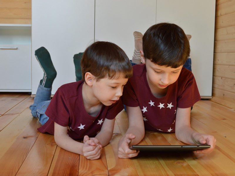 Dua anak laki-laki sedang berbaring di lantai kayu sambil memainkan tablet.