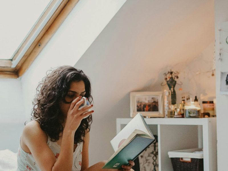 Pagi yang Nyaman dengan Buku dan Teh Seorang wanita duduk di tempat tidur, minum teh, dan membaca buku.