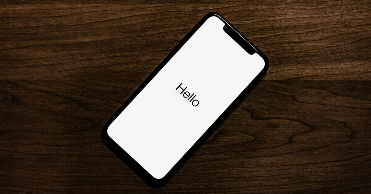 Sebuah Iphone berwarna hitam yang menampilkan kata "Halo" pada layar putih diposisikan secara diagonal pada permukaan kayu berwarna gelap.
