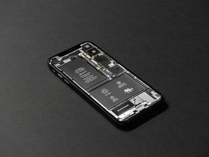 Gambar iPhone yang dibongkar dengan penutup belakang hitam dilepas, memperlihatkan komponen internal termasuk baterai, motherboard, dan komponen elektronik lainnya.