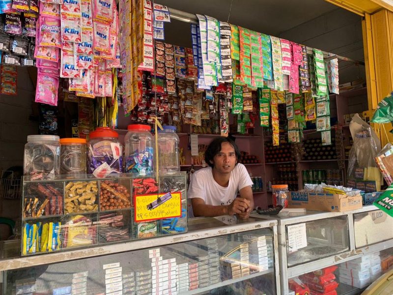 Seorang pemuda berdiri di depan etalase toko kecil yang menjual berbagai produk makanan dan minuman.