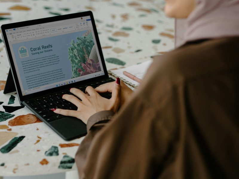 Pekerja Kreatif Menciptakan Kesadaran tentang Terumbu Karang Wanita mengenakan hijab sedang bekerja di laptop dengan tema terumbu karang di meja berwarna-warni.